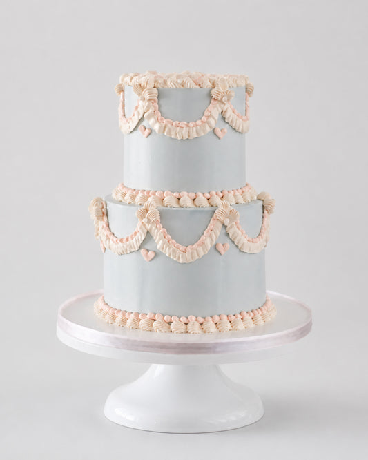 A two-tiered cake with a pale blue-gray finish, decorated with peach-pink piped garland and bow motifs, displayed on a white cake stand against a light gray background.