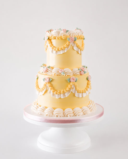 Two-tier butter yellow vintage buttercream cake with golden piped swags, white ruffle fan borders, and blush pink rosettes on a white cake stand