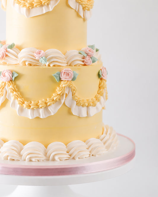 Close-up of two-tier yellow vintage buttercream cake with golden swag piping, cream ruffle fan clusters, and pink piped roses with sage green leaves