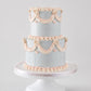 A two-tiered cake with a pale blue-gray finish, decorated with peach-pink piped garland and bow motifs, displayed on a white cake stand against a light gray background.