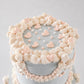 A close-up of a decorated cake with pale gray frosting, piped cream and blush pink frosting borders, and small heart decorations on top, set against a soft light gray background.