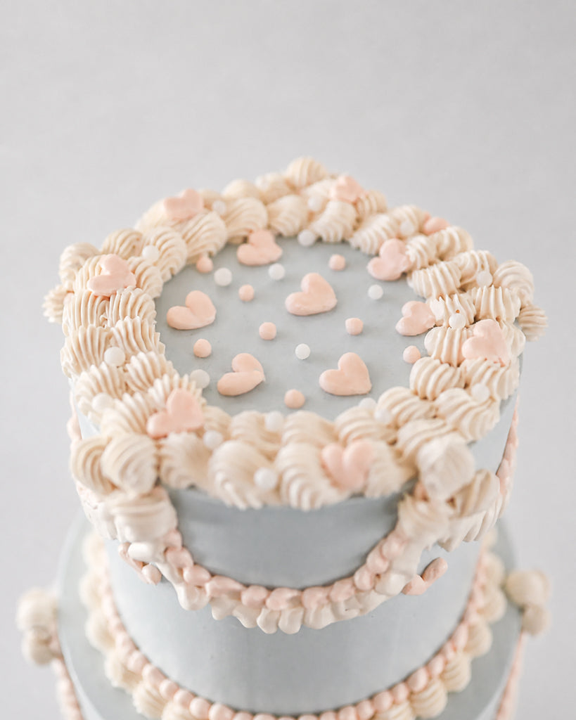 A close-up of a decorated cake with pale gray frosting, piped cream and blush pink frosting borders, and small heart decorations on top, set against a soft light gray background.