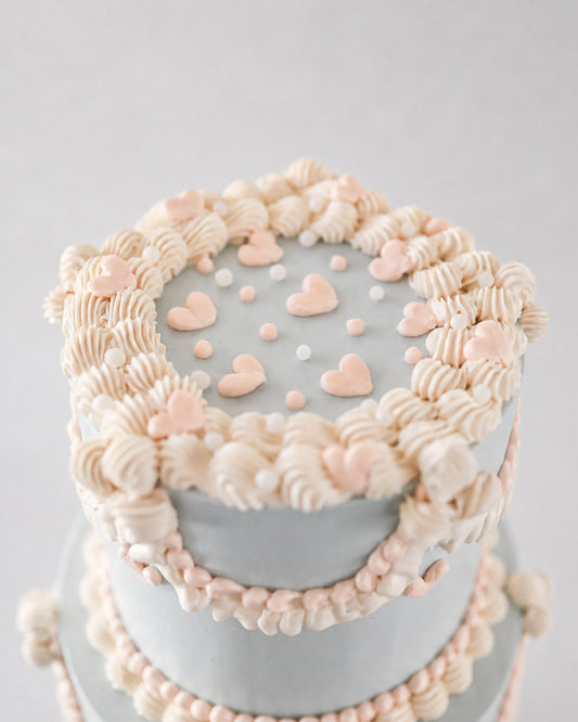 A close-up of a decorated cake with pale gray frosting, piped cream and blush pink frosting borders, and small heart decorations on top, set against a soft light gray background.