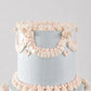 A close-up of a two-tier cake with smooth light blue frosting and delicate pale pink piped decorations, including ruffles, garlands, and small hearts, against a soft light gray background.