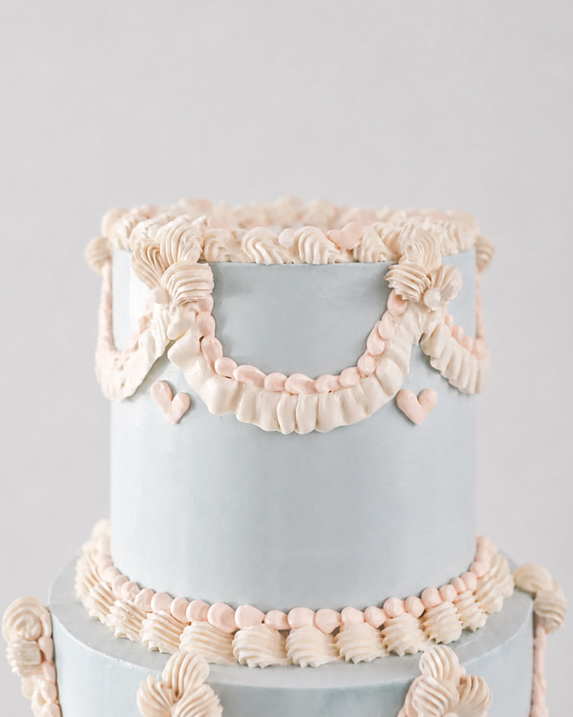 A close-up of a two-tier cake with smooth light blue frosting and delicate pale pink piped decorations, including ruffles, garlands, and small hearts, against a soft light gray background.