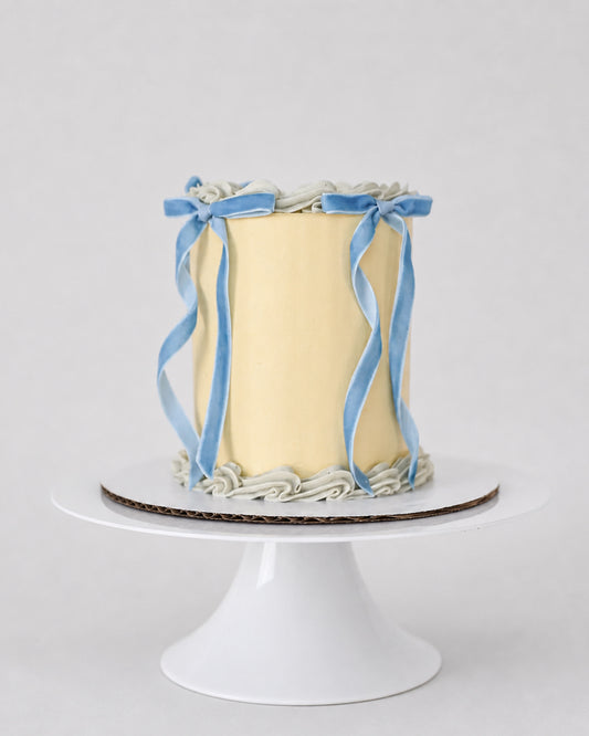 Bluebell Bow mini cake in soft yellow with pale blue shell piping and blue velvet ribbon bows