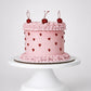Blush pink coquette cake with dark red piped heart dots, pink rosette scroll border, and three glossy red cherries with stems on a white cake stand