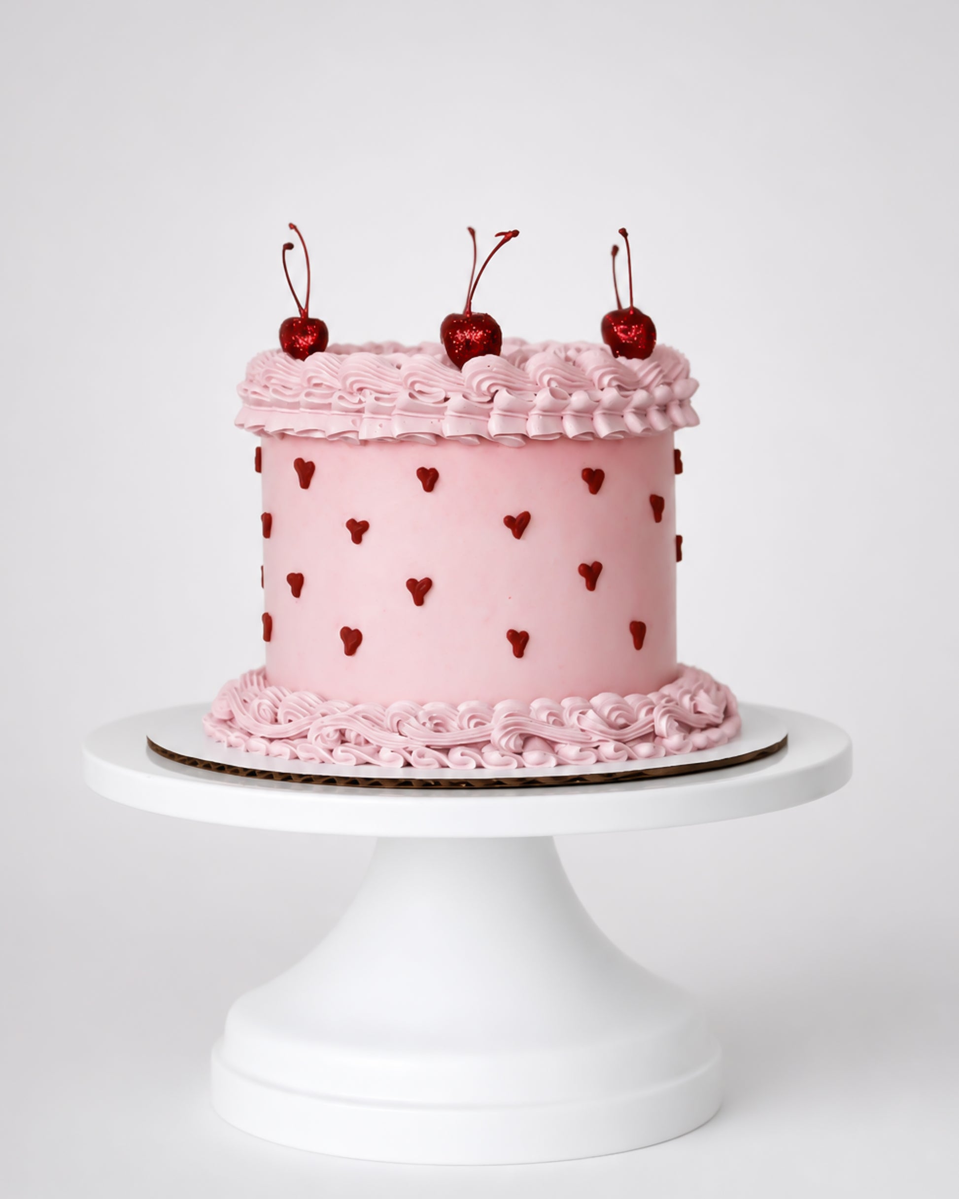 Blush pink coquette cake with dark red piped heart dots, pink rosette scroll border, and three glossy red cherries with stems on a white cake stand