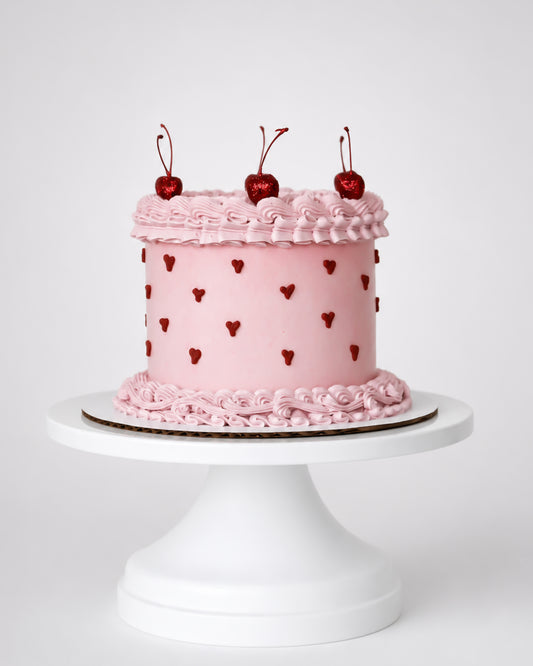 Blush pink coquette cake with dark red piped heart dots, pink rosette scroll border, and three glossy red cherries with stems on a white cake stand