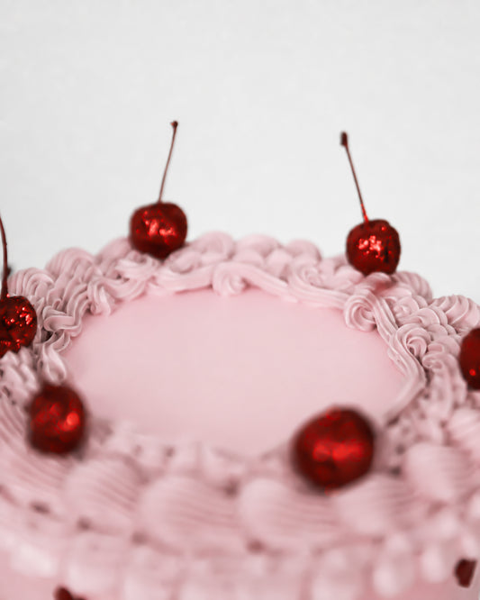 Top view of blush pink coquette cake with piped pink rosette border and glossy lacquered red cherries with stems