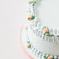 Angled close-up of Gail Marie cake showing smooth mint green sides with pearl dot details and hand-piped ruffle borders accented with peach rosettes and sage leaves