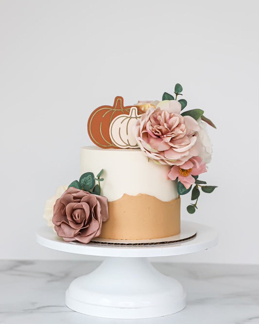 A decorated cake with acrylic pumpkin topper and non-toxic faux florals, placed on a white cake stand against a neutral background.