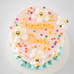 Top view of blush pink birthday cake with hand-piped happy Day message in golden buttercream, rainbow sprinkles, and smiley daisy flowers