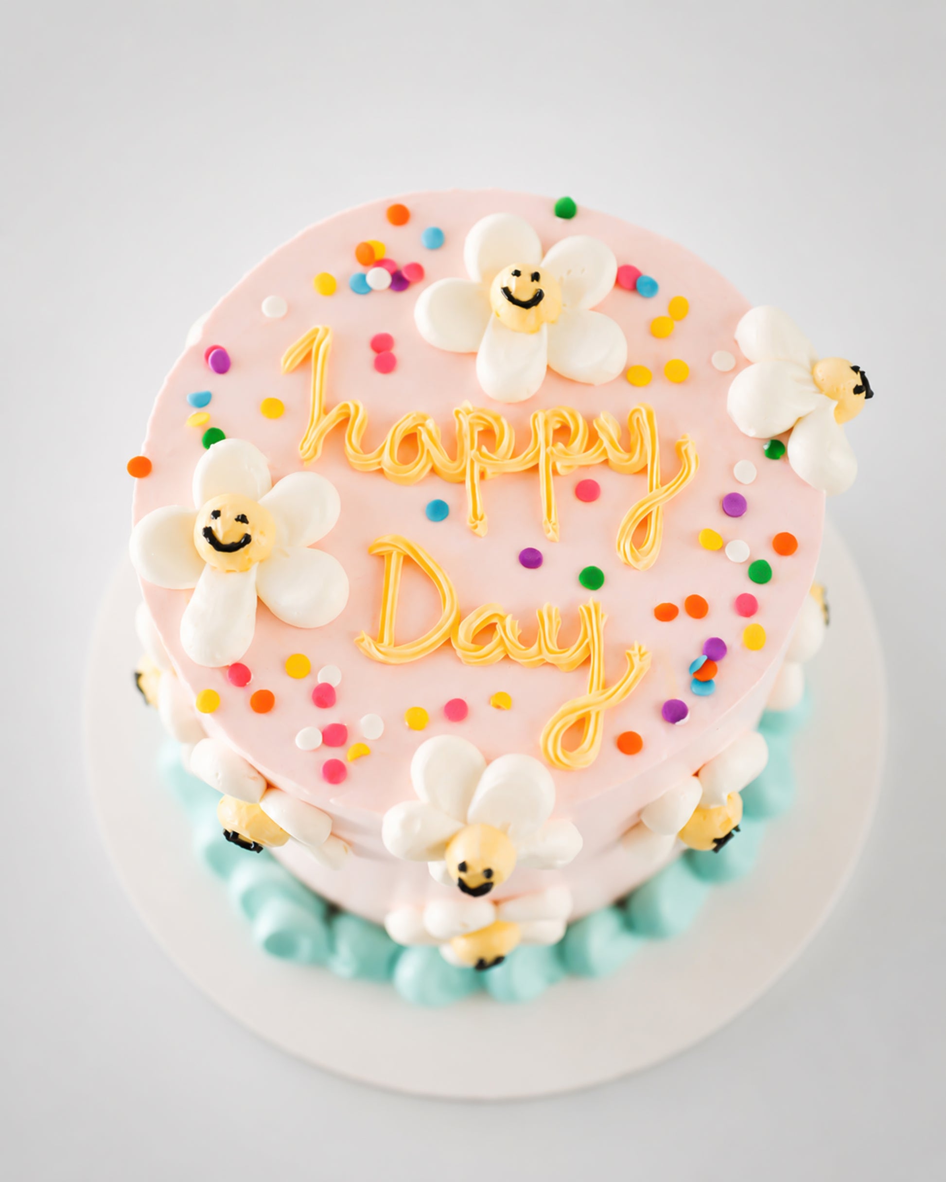 Top view of blush pink birthday cake with hand-piped happy Day message in golden buttercream, rainbow sprinkles, and smiley daisy flowers