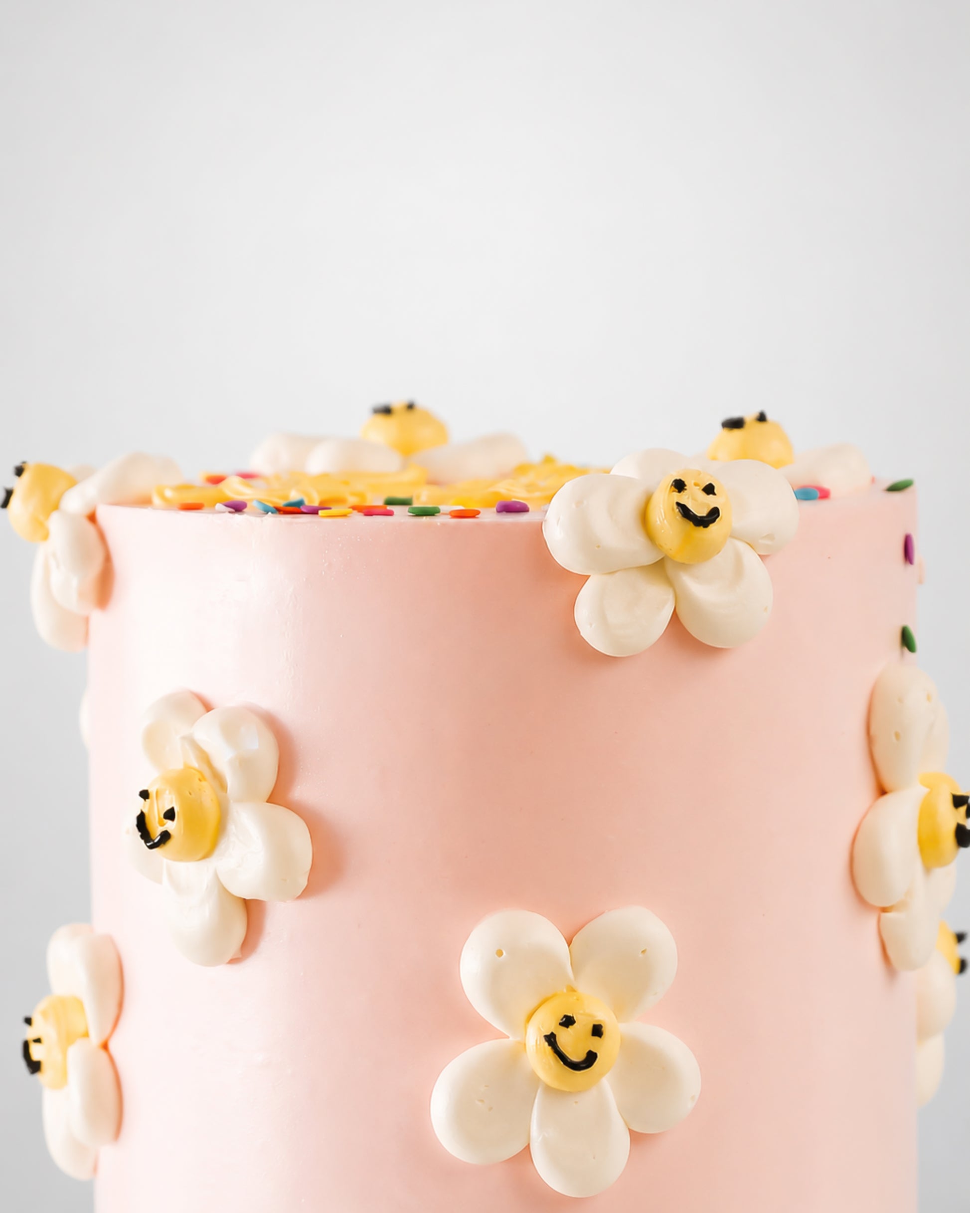 Close-up of blush pink buttercream cake with scattered white piped daisy flowers with yellow smiley face centers
