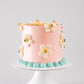 Blush pink vintage birthday cake with white smiley daisy flowers and teal shell border on a white cake stand