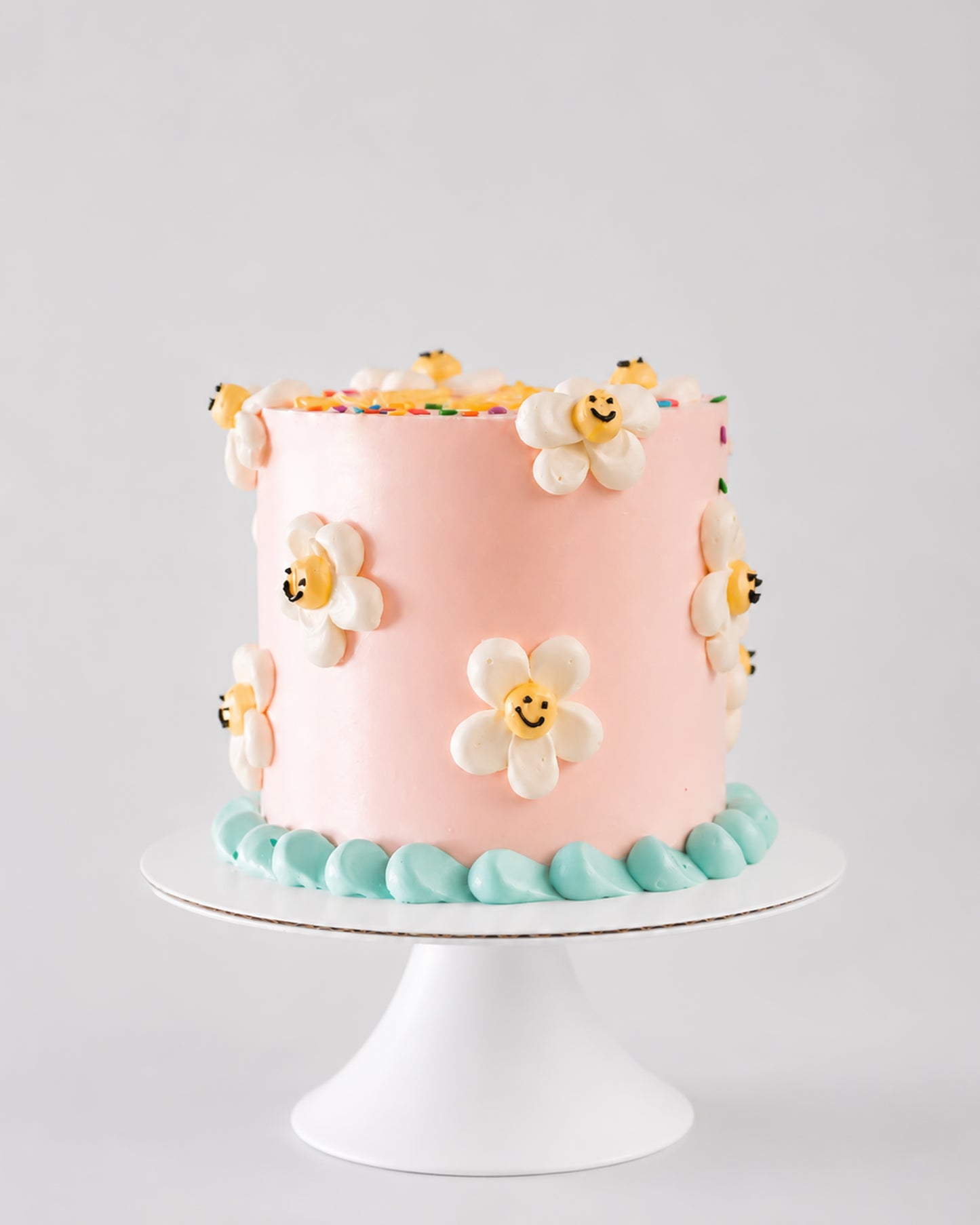 Blush pink vintage birthday cake with white smiley daisy flowers and teal shell border on a white cake stand