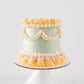 Mary Lou cake with sage green buttercream, white ruffle swag border with pink bow accents, and a crown of pale yellow rosettes