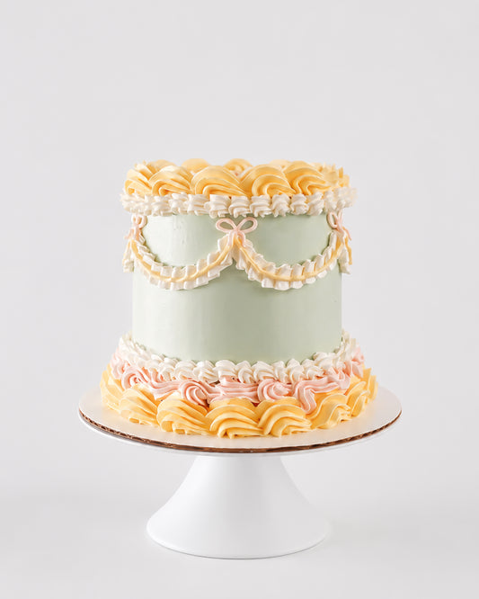 Mary Lou cake with sage green buttercream, white ruffle swag border with pink bow accents, and a crown of pale yellow rosettes