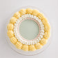Top-down view of Mary Lou cake showing smooth sage green center surrounded by white scroll piping with pink pearl beads and an outer ring of pale yellow rosettes