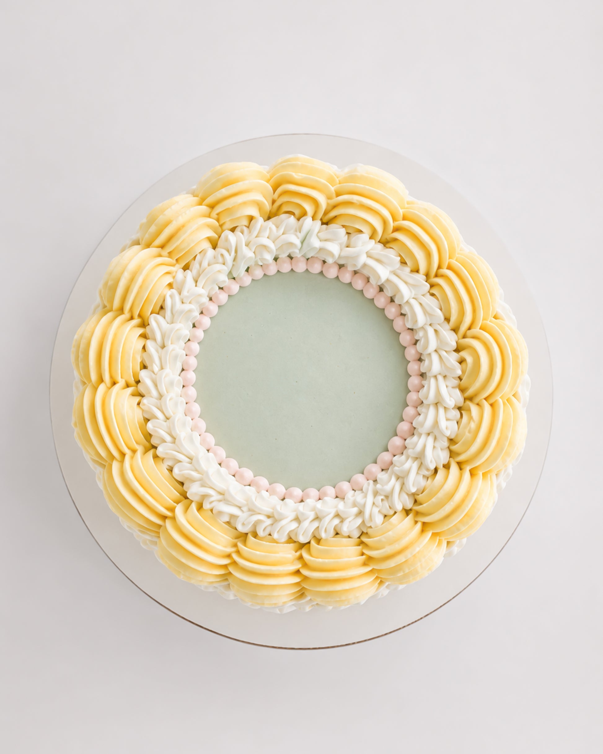Top-down view of Mary Lou cake showing smooth sage green center surrounded by white scroll piping with pink pearl beads and an outer ring of pale yellow rosettes
