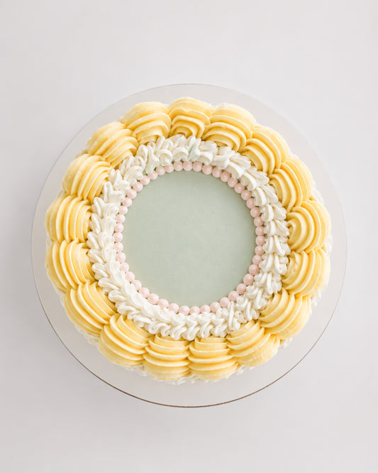 Top-down view of Mary Lou cake showing smooth sage green center surrounded by white scroll piping with pink pearl beads and an outer ring of pale yellow rosettes