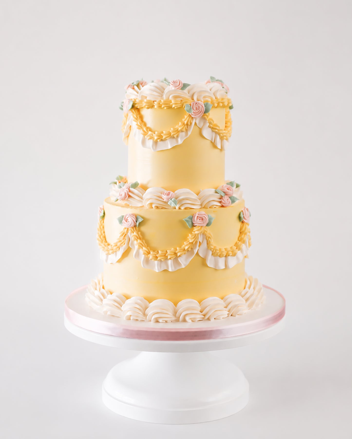 Two-tier butter yellow vintage buttercream cake with golden piped swags, white ruffle fan borders, and blush pink rosettes on a white cake stand