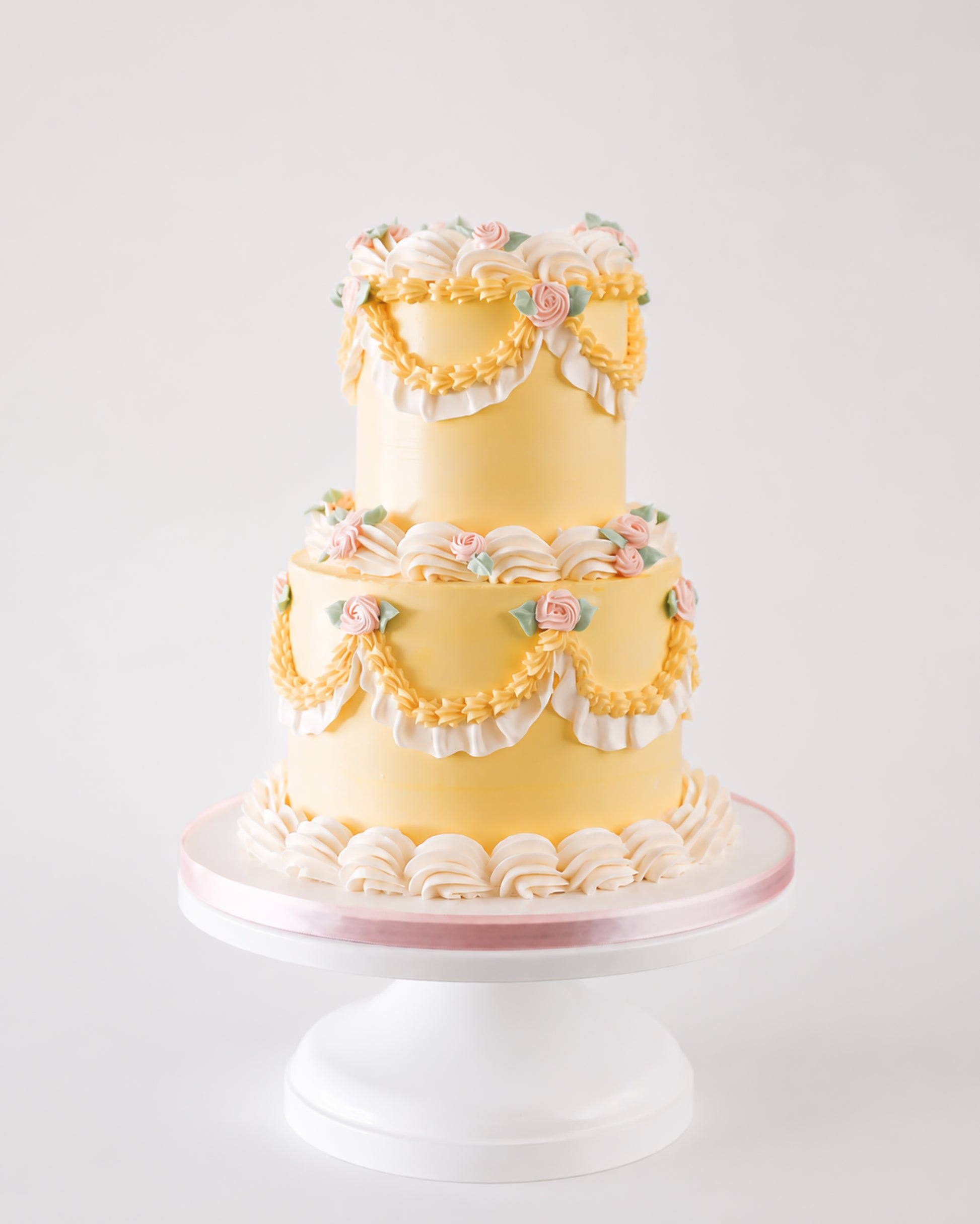 Two-tier butter yellow vintage buttercream cake with golden piped swags, white ruffle fan borders, and blush pink rosettes on a white cake stand