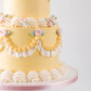 Close-up of two-tier yellow vintage buttercream cake with golden swag piping, cream ruffle fan clusters, and pink piped roses with sage green leaves