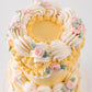 Top-down view of two-tier vintage yellow buttercream cake with crown of cream ruffle fans and blush pink piped rosettes with sage leaves
