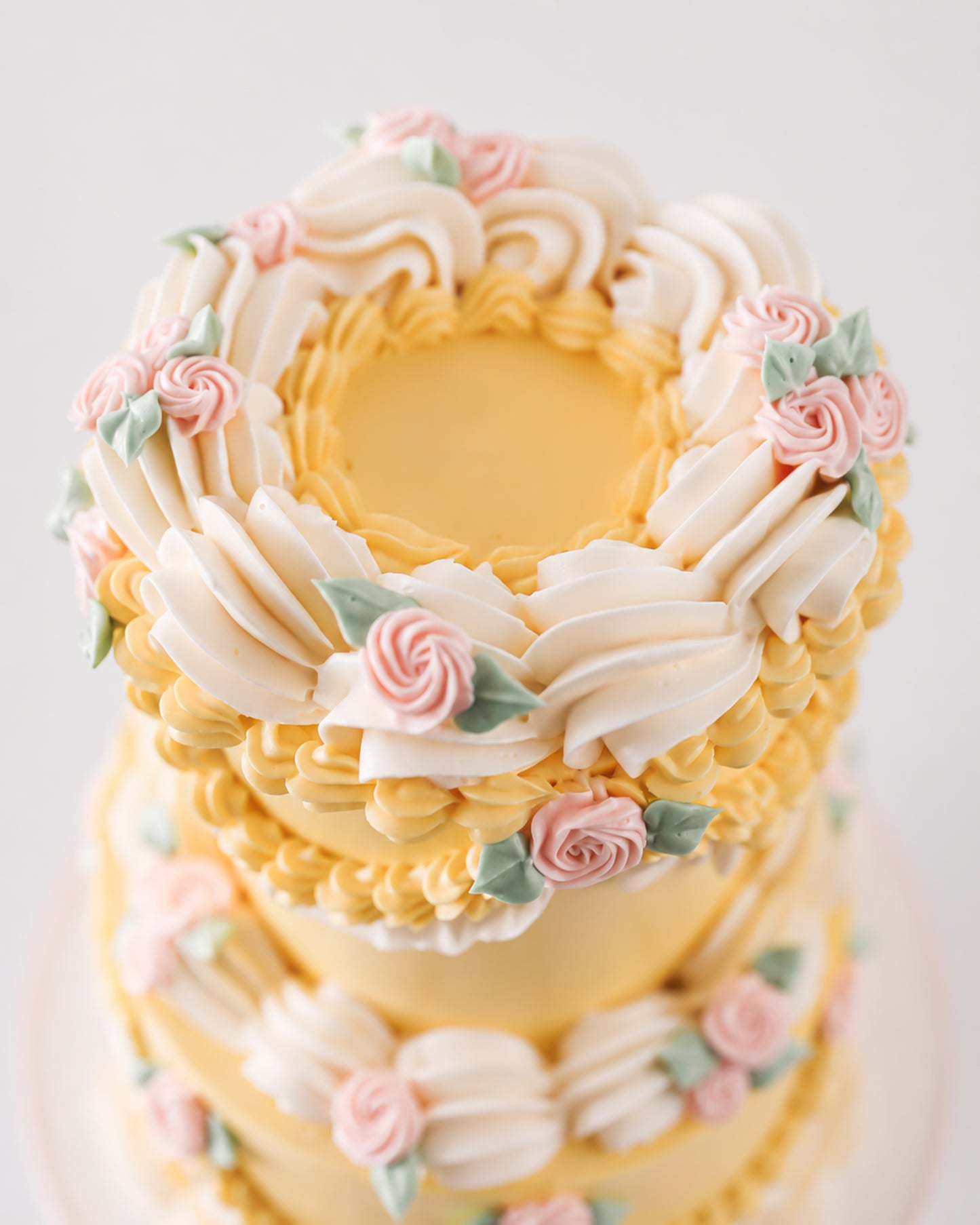 Top-down view of two-tier vintage yellow buttercream cake with crown of cream ruffle fans and blush pink piped rosettes with sage leaves