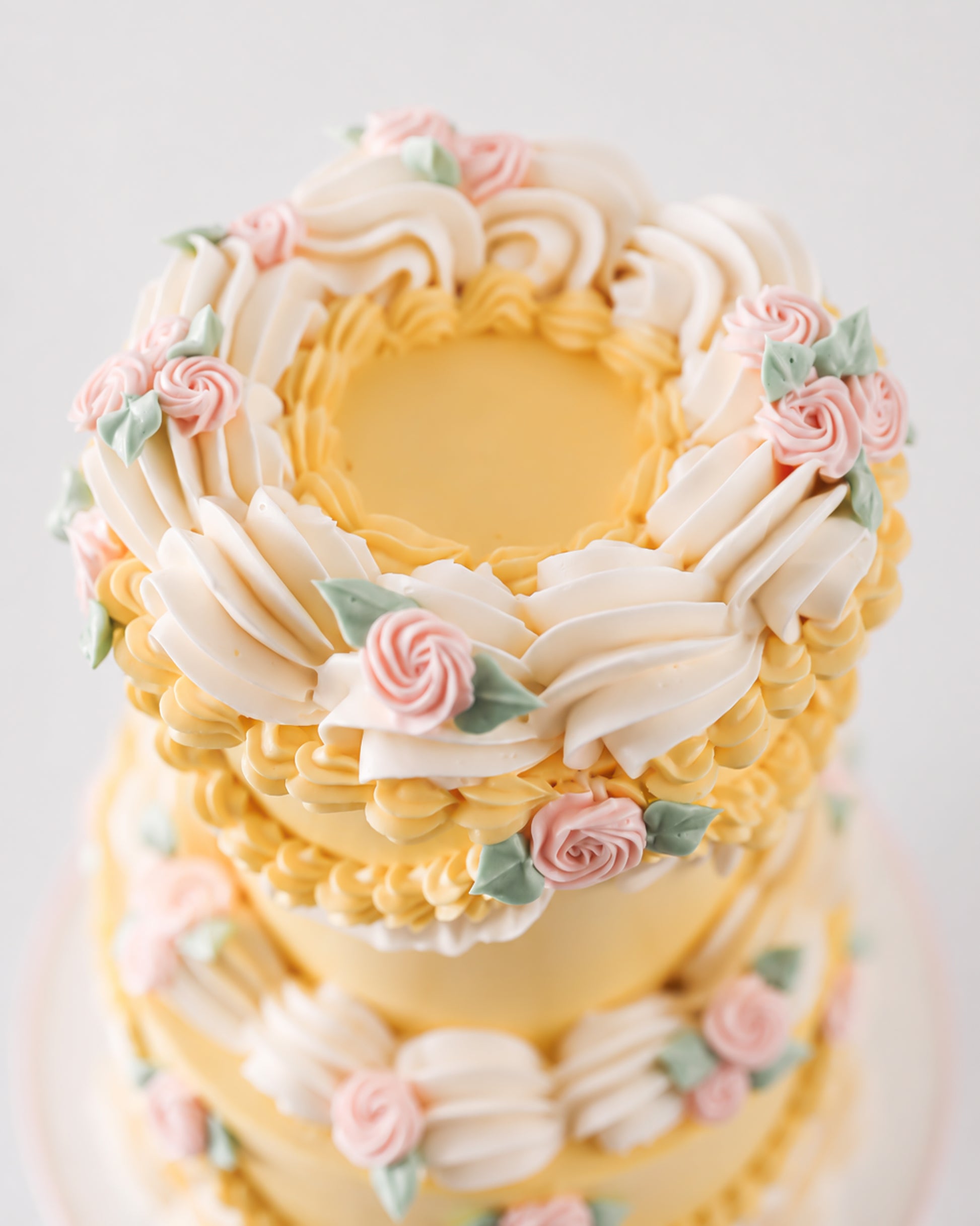 Top-down view of two-tier vintage yellow buttercream cake with crown of cream ruffle fans and blush pink piped rosettes with sage leaves